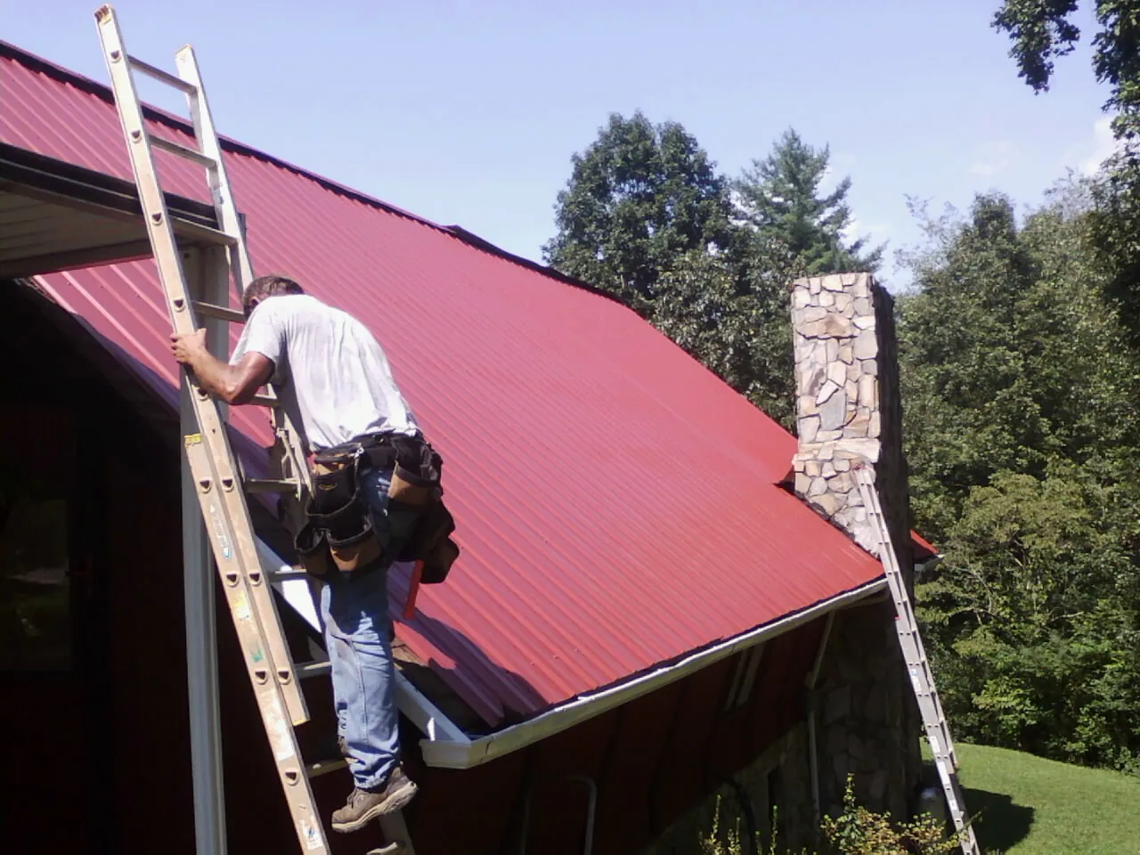 Roofer on a ladder beside a red metal roof and stone chimney — Soffit & Fascia Repair by Roofing Brothers in Early Branch